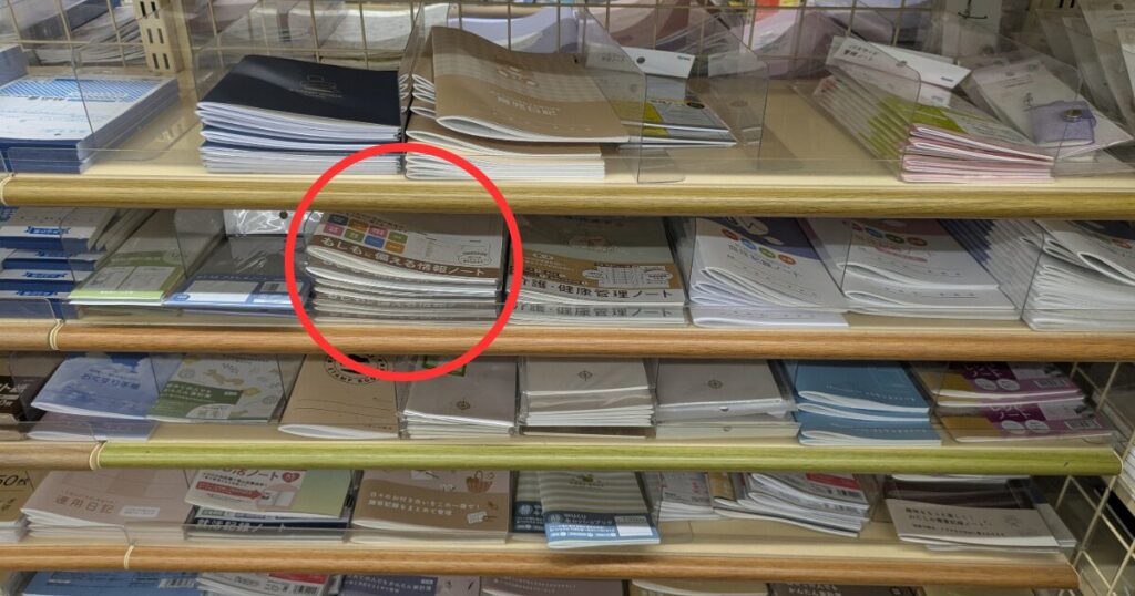 00円ショップのセリア店舗にある文具売り場の棚。赤丸で囲まれた位置に、薄型で白い表紙のエンディングノート「もしもに備える情報ノート」が数冊積まれて陳列されている。周囲には他の実用ノートやお薬手帳なども並んでいる。